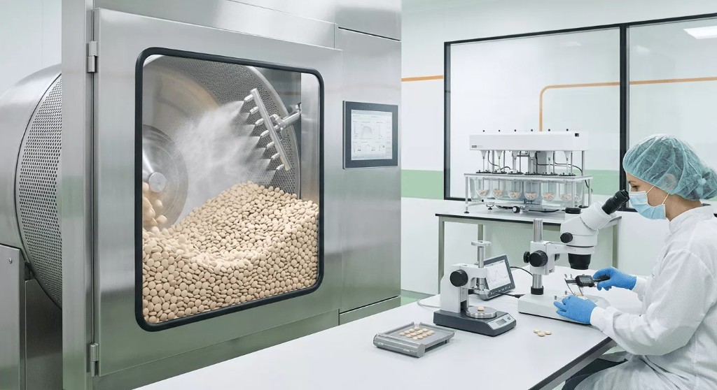 Tablet coating machine tumbling tablets beside a QC workstation with microscope and hardness tester